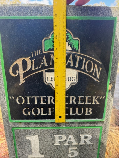 The same hole-marker monument being measured on site with a tape measure, documenting the exact dimensions needed to fabricate a QR-enabled sponsor plaque insert that would fit the existing stone.